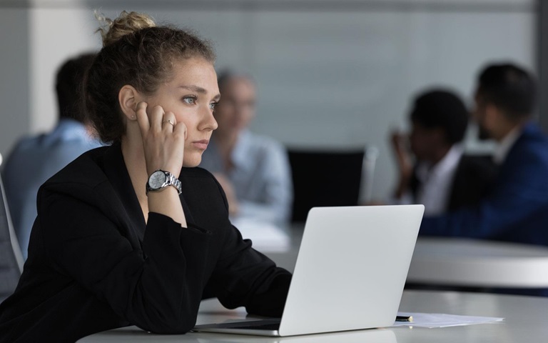 Young thoughtful woman sitting at desk with laptop looking into distance feeling unmotivated