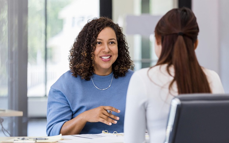 adult woman and young adult female co-worker sit across from each other to brainstorm for the project