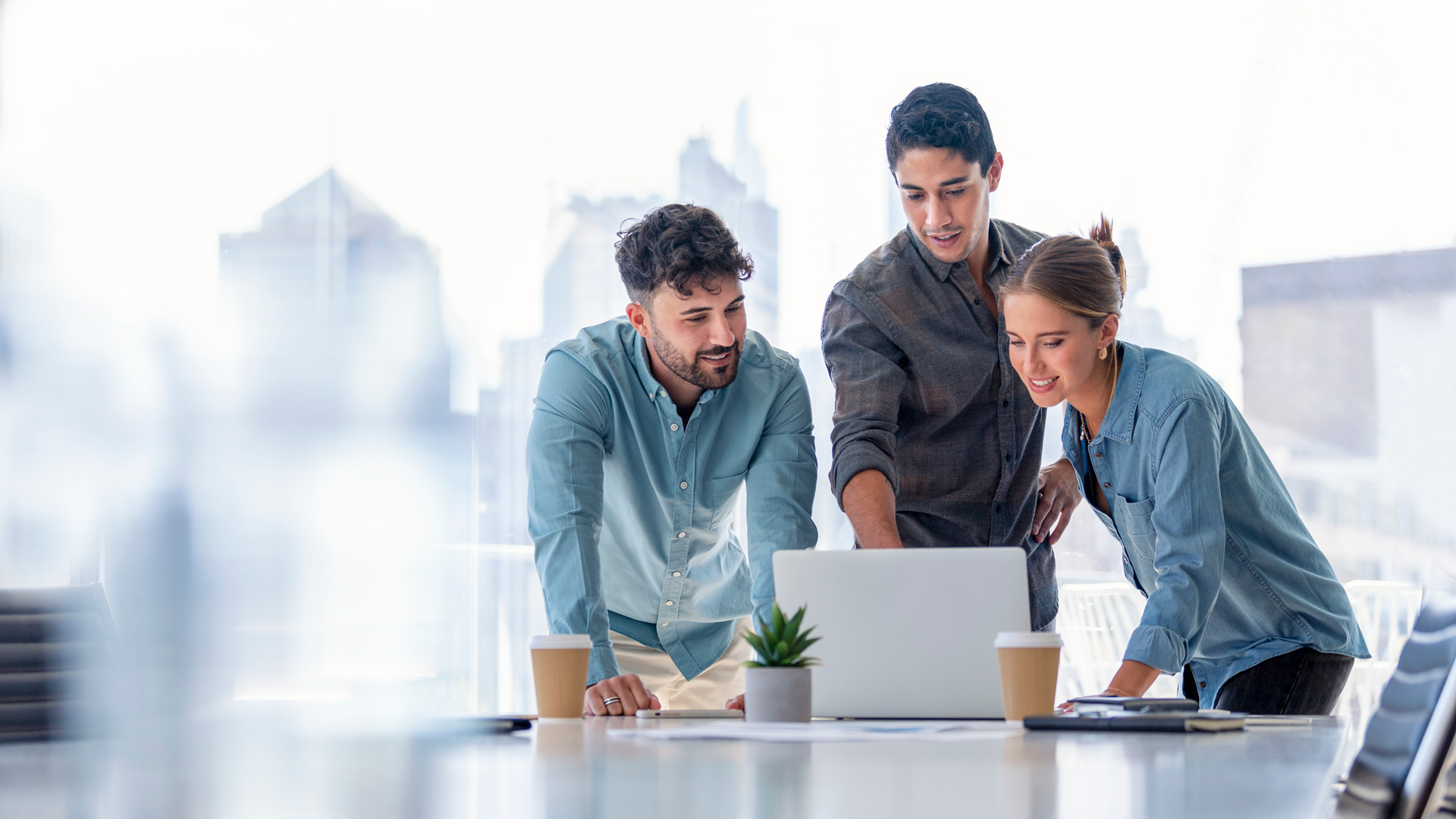 three employees looking at laptop computer in open office space with large windows